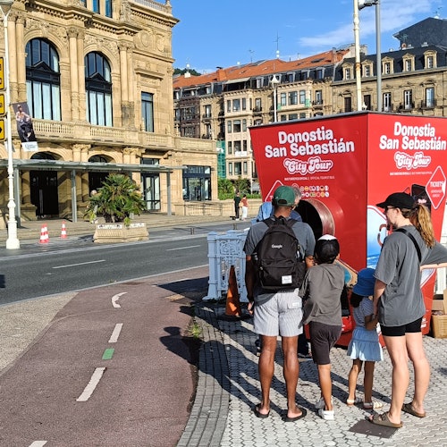 A family examines a red Donostia San Sebastian City Tour kiosk on a sunny street near historic buildings.