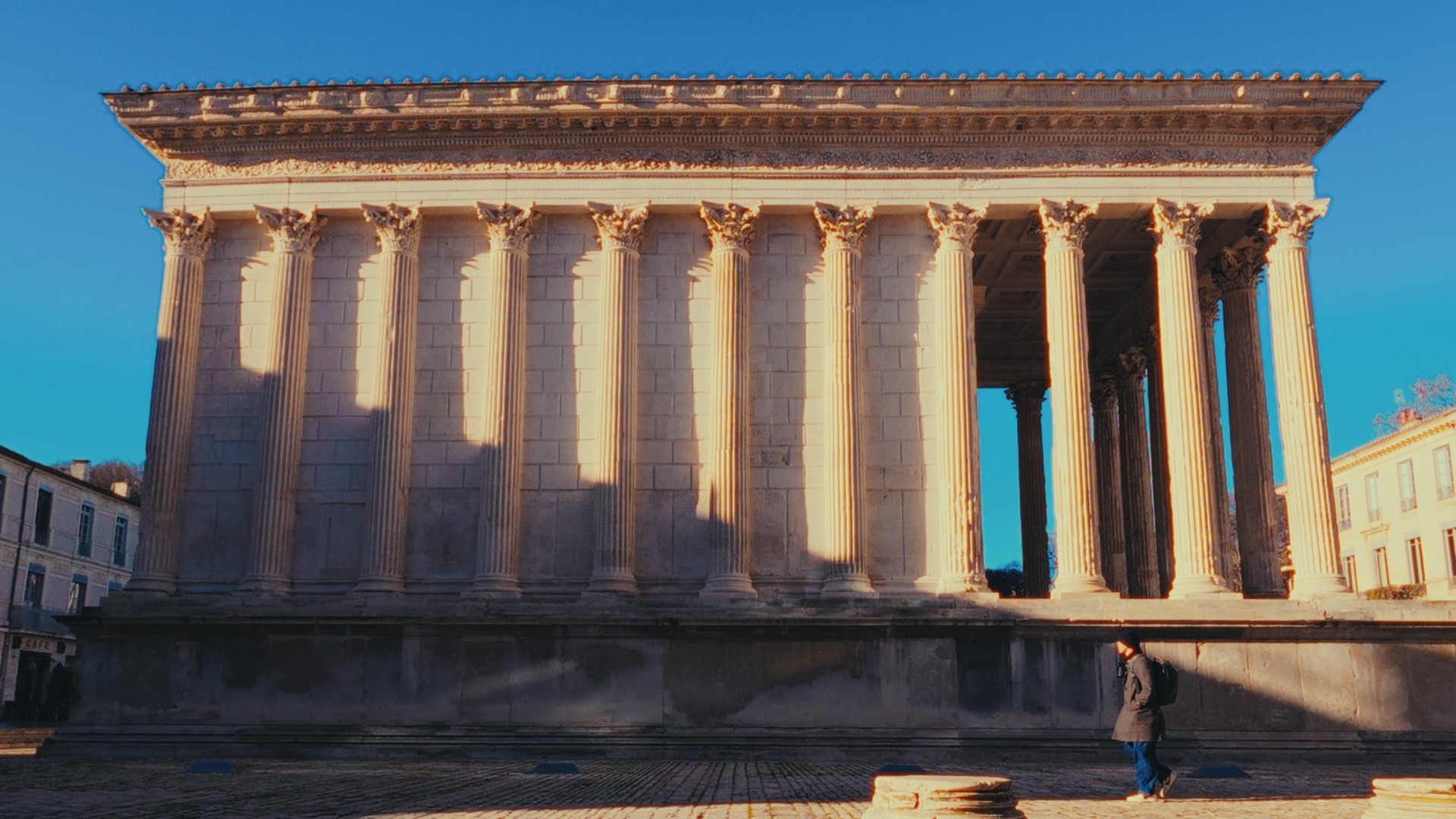 Silhouette d’une personne marchant devant la Maison Carrée, sous une lumière dorée du matin.