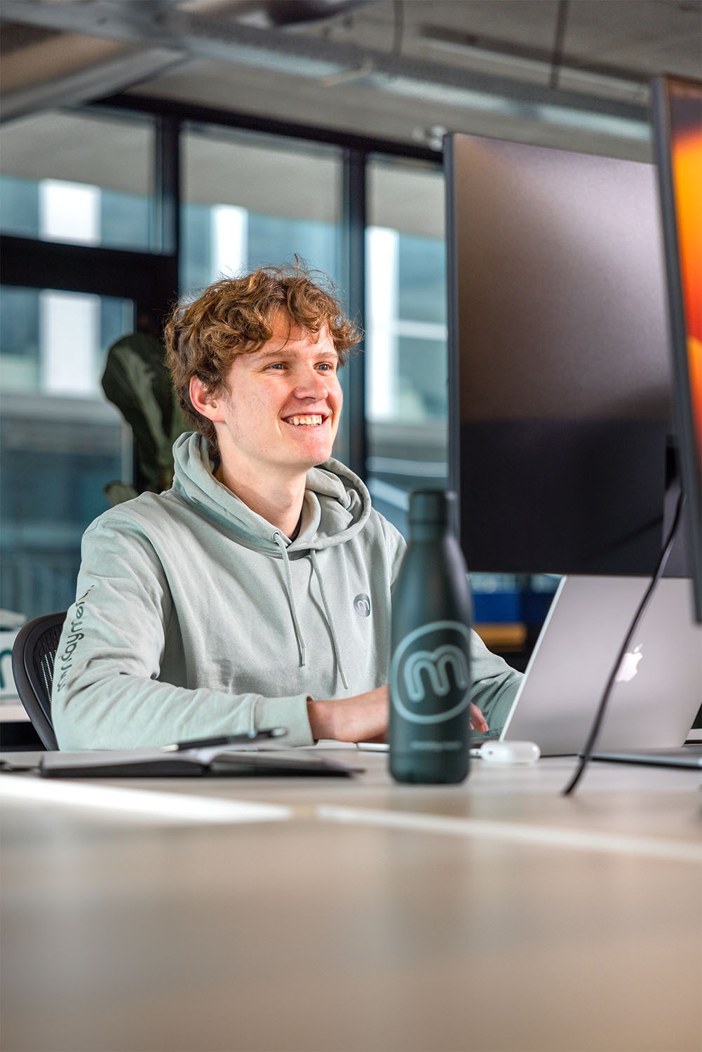 A person in a hoodie smiles at a computer screen in an office setting, with a branded water bottle nearby