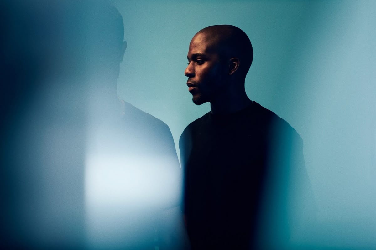 Portrait of a Black man with a shaved head in profile, looking contemplative against a blue backdrop with blurred foreground.