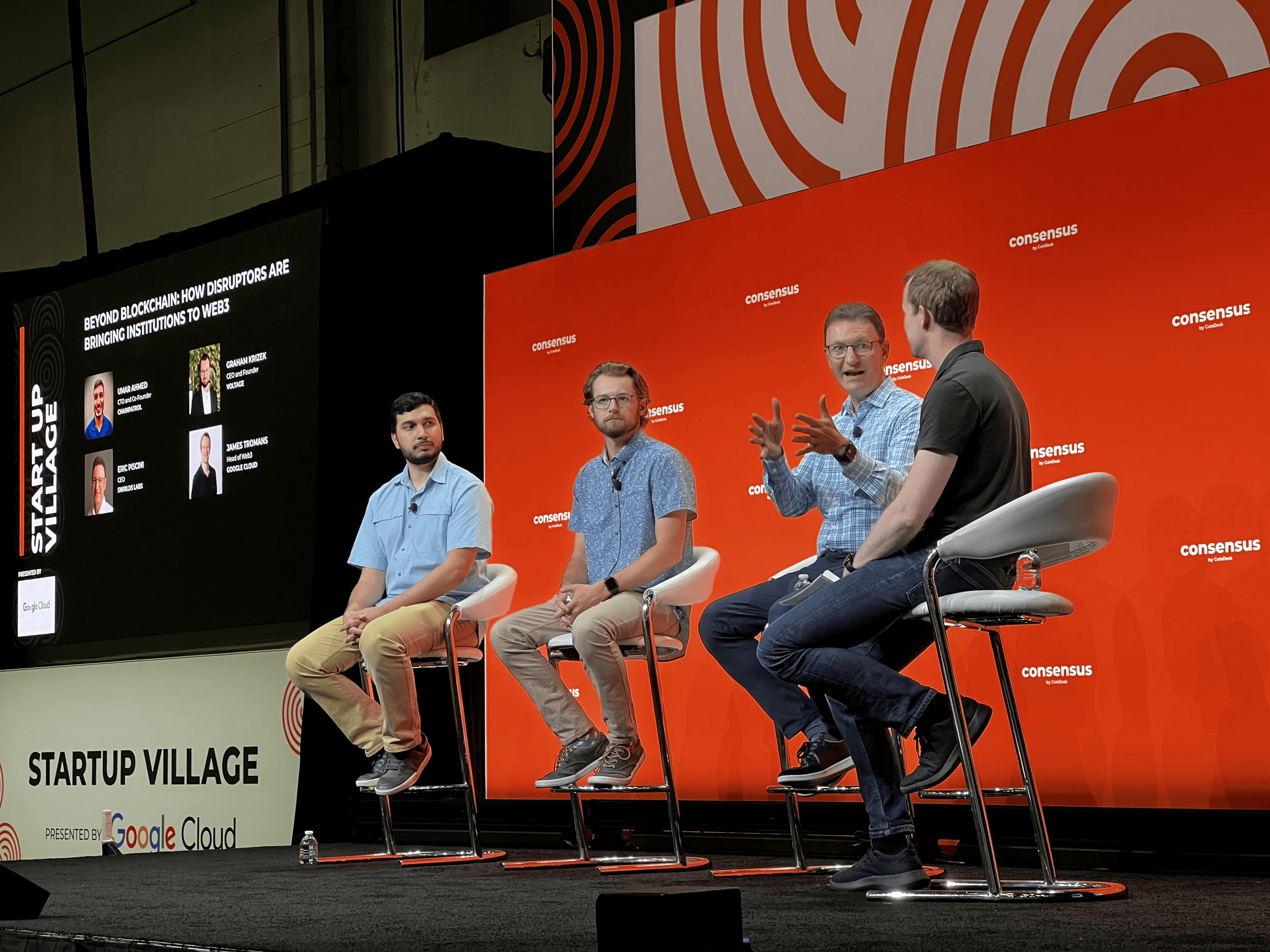 4 men sitting on chairs on a stage, for a panel at Consensus, with a red background behind them