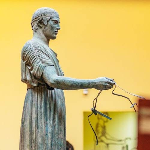 Estátua antiga de bronze de uma figura em pé, segurando objetos finos e delicados, diante de um fundo amarelo em um ambiente interno de museu.