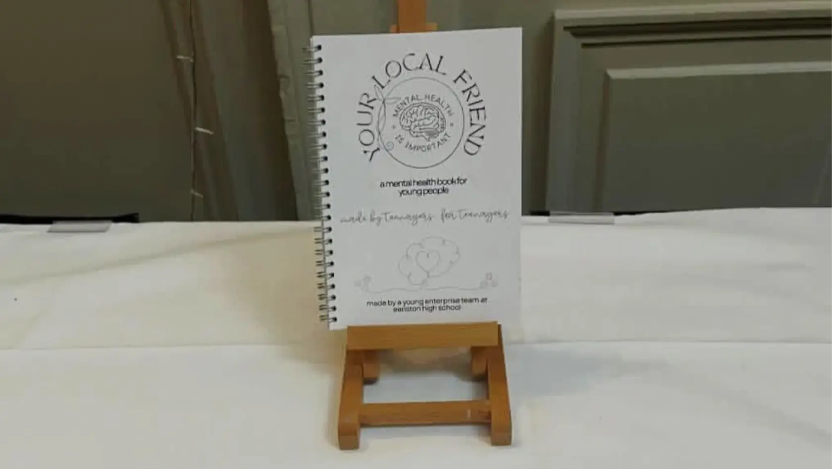 A spiral-bound mental health book titled "Your Local Friend" is displayed on a small wooden easel against a neutral background, conveying a supportive tone.