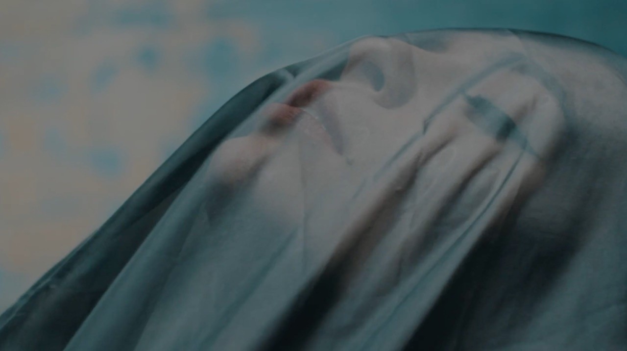 Close-up of a person’s face seen through a sheer dark veil, features blurred and softly lit.