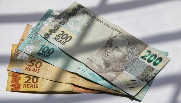Cédulas de real brasileiro (R$20, R$50, R$100 e R$200) sobre superfície iluminada e fundo branco.