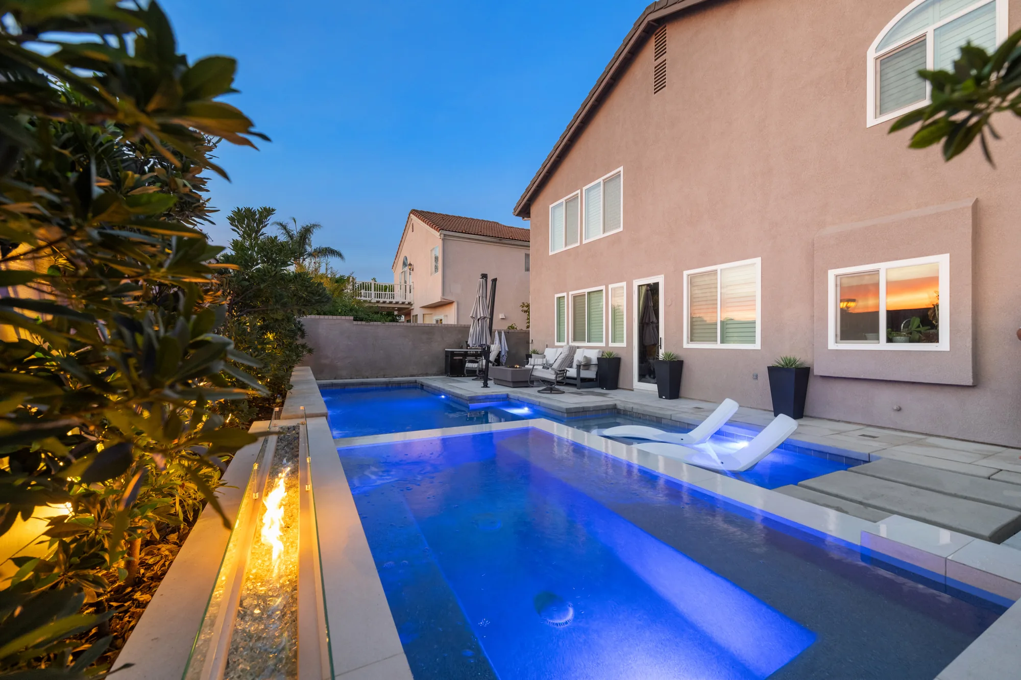 Paved pool edge with white loungers beside a home and a fire feature across the water.