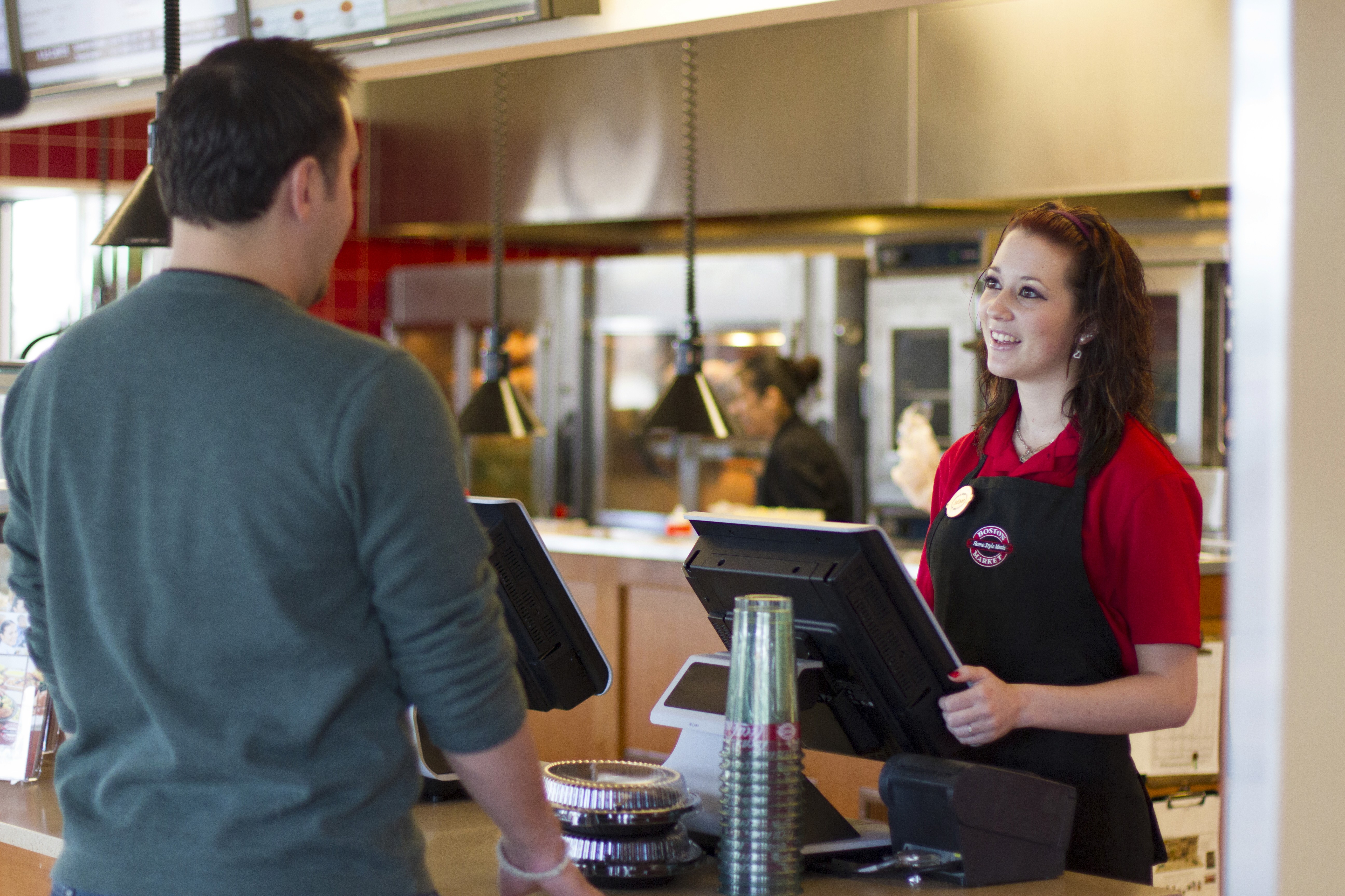 A thick cut of steak with prominent grill marks sizzles over hot coals and flames on a barbecue, with blurred figures of people and drinks in the background, suggesting an outdoor gathering.