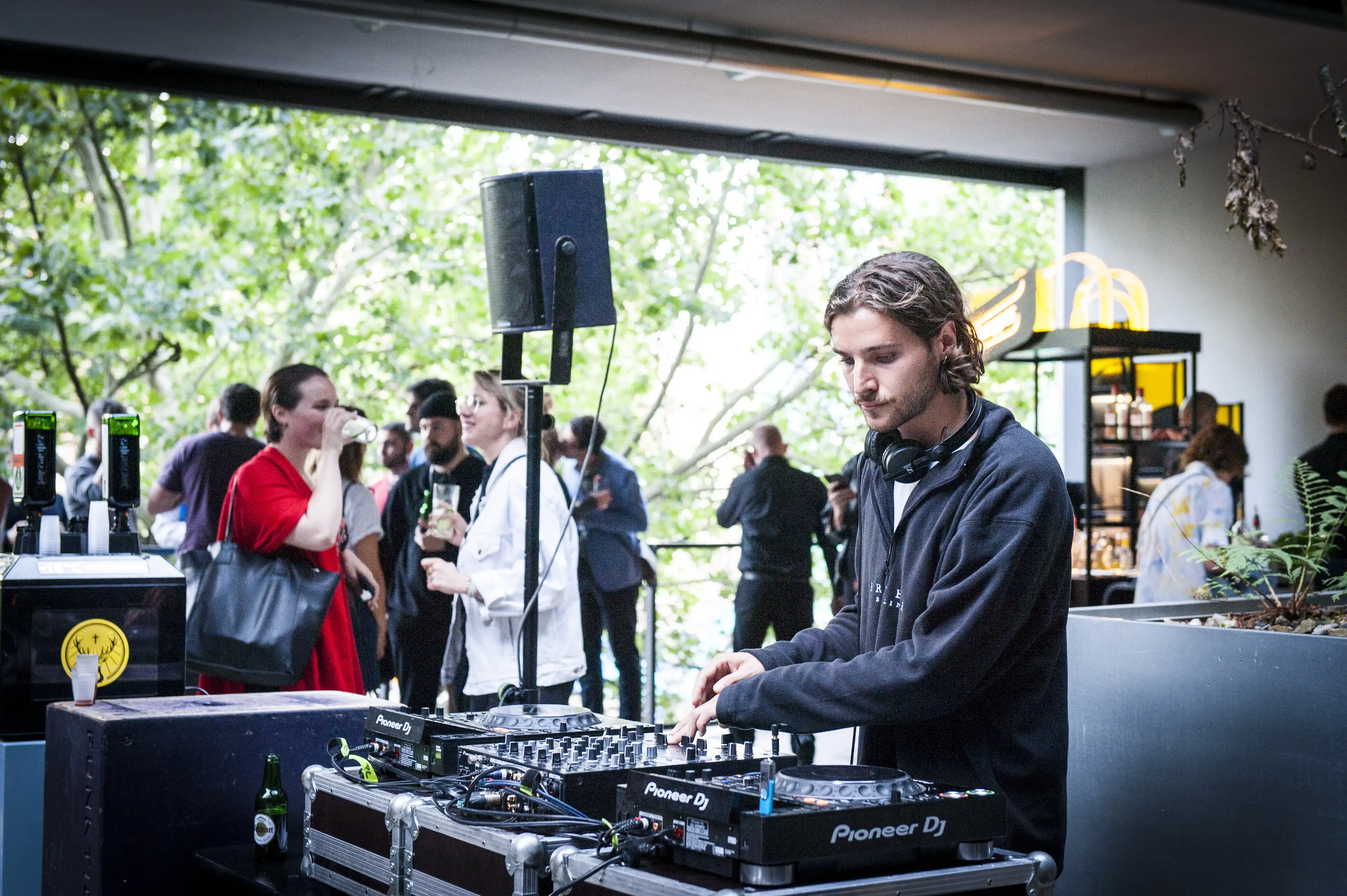 DJ playing on Pioneer decks at the STUDIOLO opening event with guests holding drinks in the green courtyard behind.
