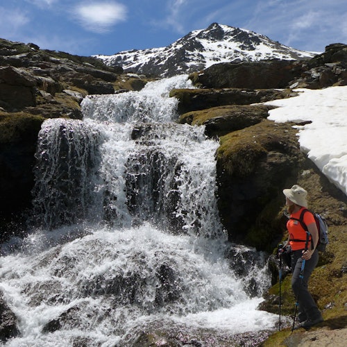 一位穿红色衬衫的徒步旅行者站在蓝天下的岩石和积雪覆盖的山上，旁边是一座瀑布。
