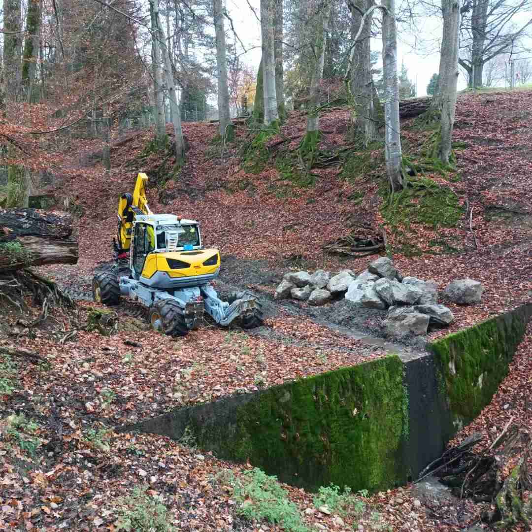 Specialized excavator working in a forest area on riverbed reinforcement and erosion control – hydraulic engineering project by Strub Bau AG.