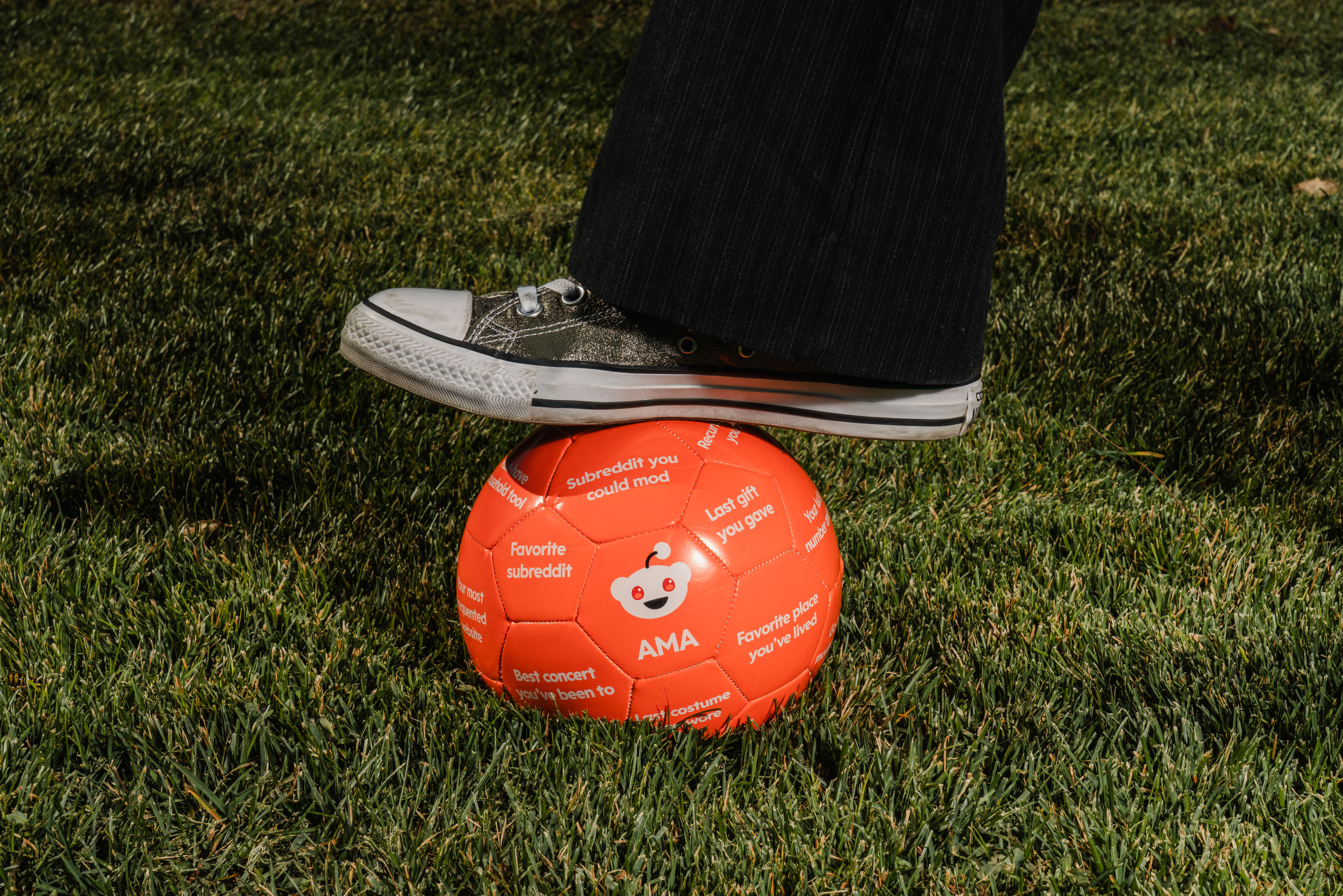 A sneaker on top of an orange soccer ball with a reddit logo on grass.