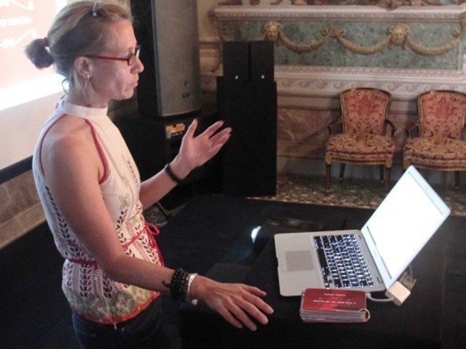 A photo of a person giving a lecture and reading from a laptop screen.