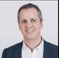 Blonde woman in a black long-sleeve and apron, arms crossed—minimalist studio portrait on a beige background.