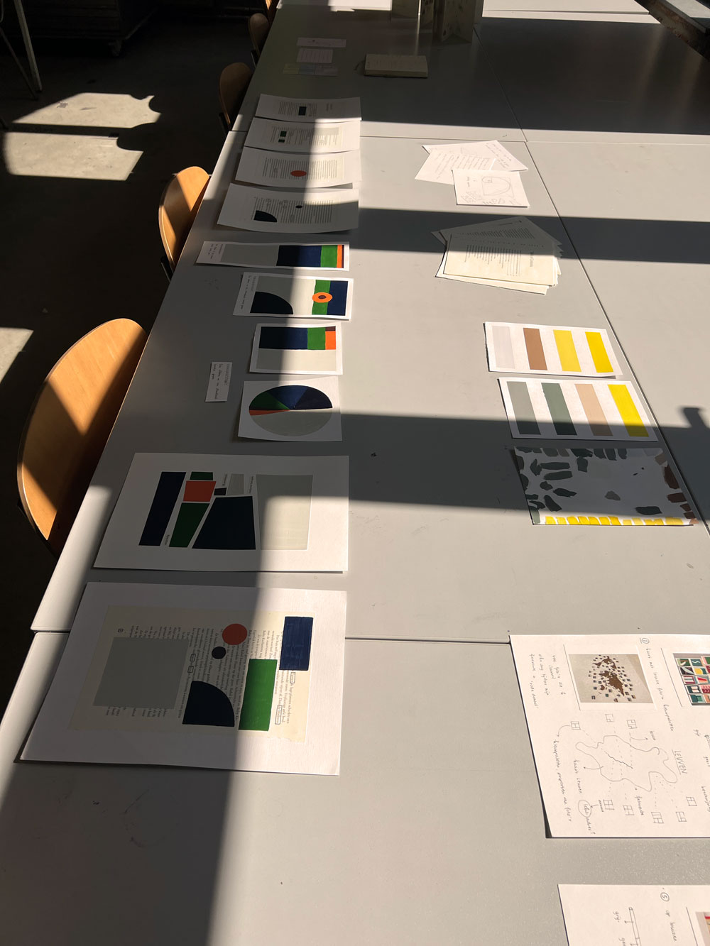 Een overzichtsfoto van een lange witte werktafel in een zonovergoten studio. Op de tafel liggen verschillende fasen van een grafisch project: vellen papier met tekst en abstracte kleurvlakken, kleurpaletten in stroken en handgetekende schema's. Harde schaduwen van ramen vallen diagonaal over het werkblad.