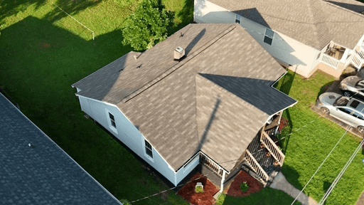 Aerial view of a single-story house with a gray roof, surrounded by green grass and nearby structures.