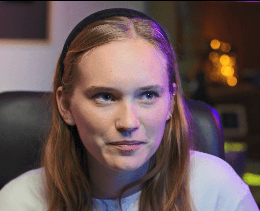 A woman with long hair stands, looking thoughtfully into the distance.