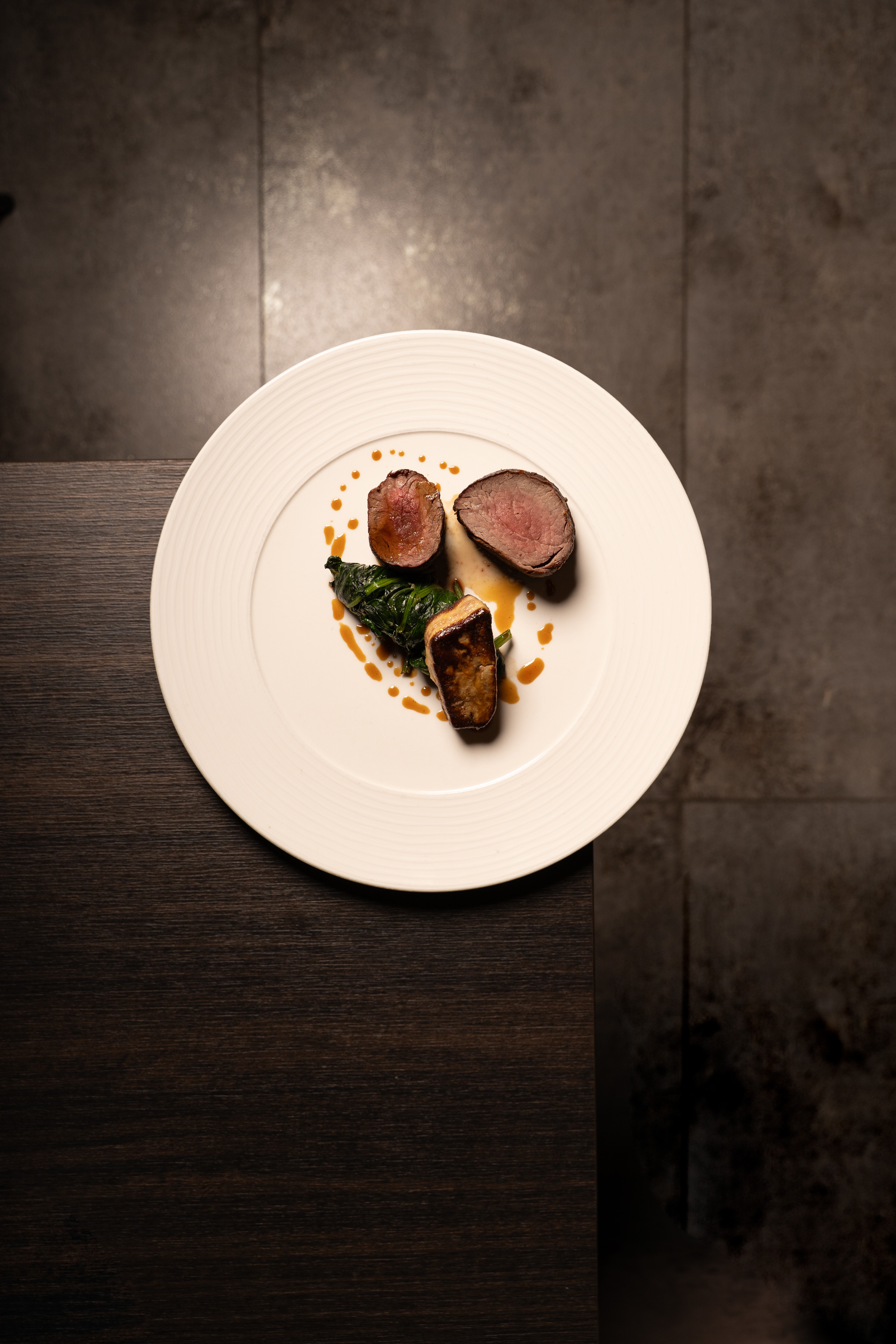 „Fein angerichtetes Rinderfilet mit sautiertem Gemüse und Jus, elegant serviert auf einem weißen Teller auf dunklem Holztisch.“