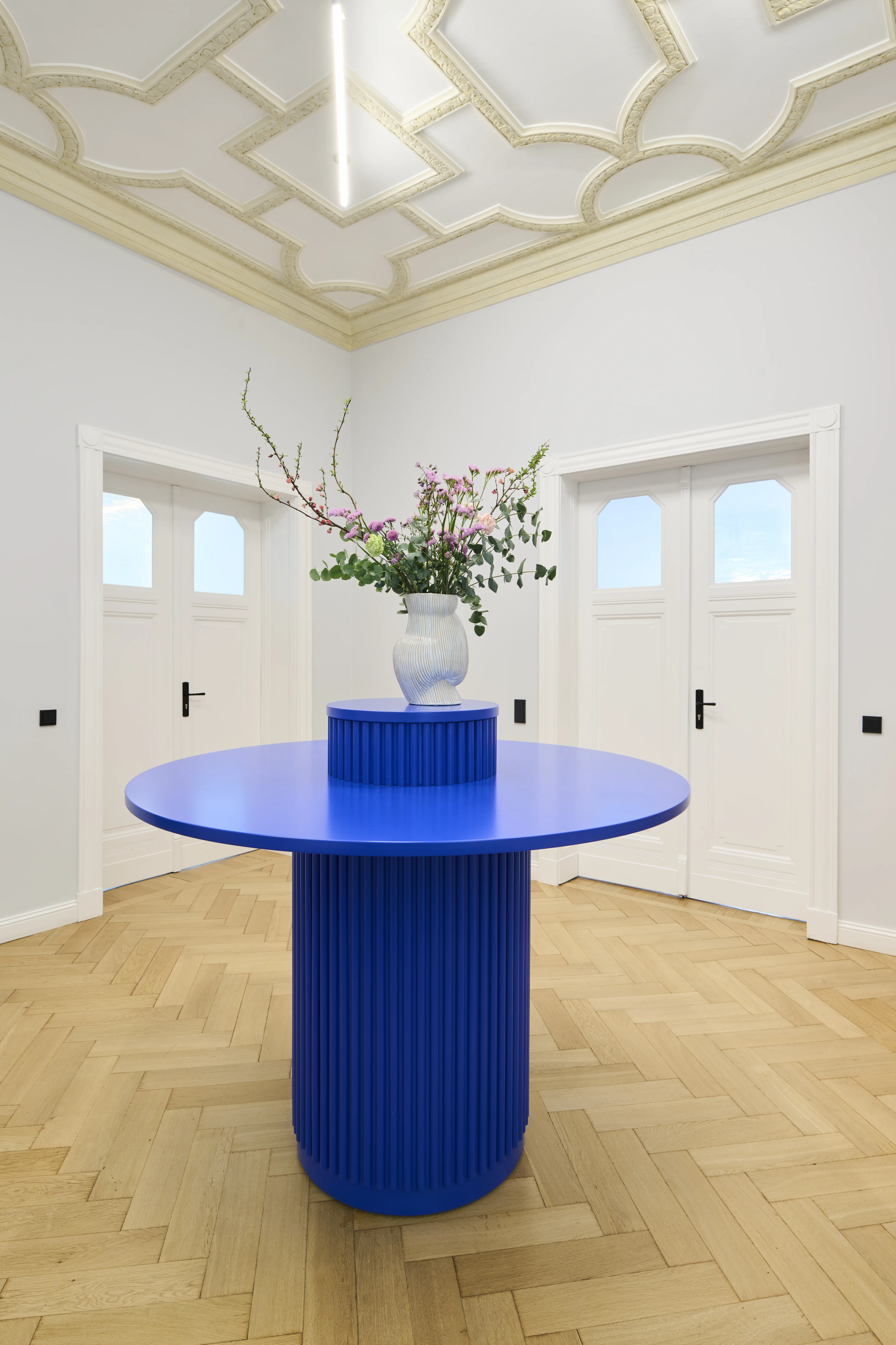 Round cobalt-blue table with ribbed pedestal base and a white vase of flowering branches at INTERMATE BERLIN, centred in a room with ornate geometric stucco ceiling and white double doors.