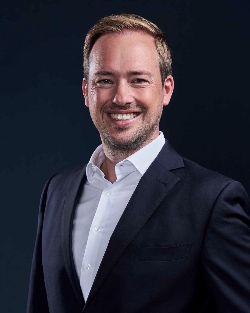 A smiling man with glasses, curly hair, wearing a maroon hoodie, poses against a dark background.