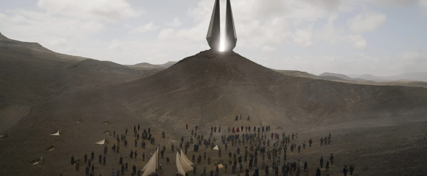 A large crowd gathers in a barren desert landscape around a tall, metallic, pyramid-like structure atop a hill. The sky is partly cloudy, and tents are scattered across the ground. The structure emits a bright light.