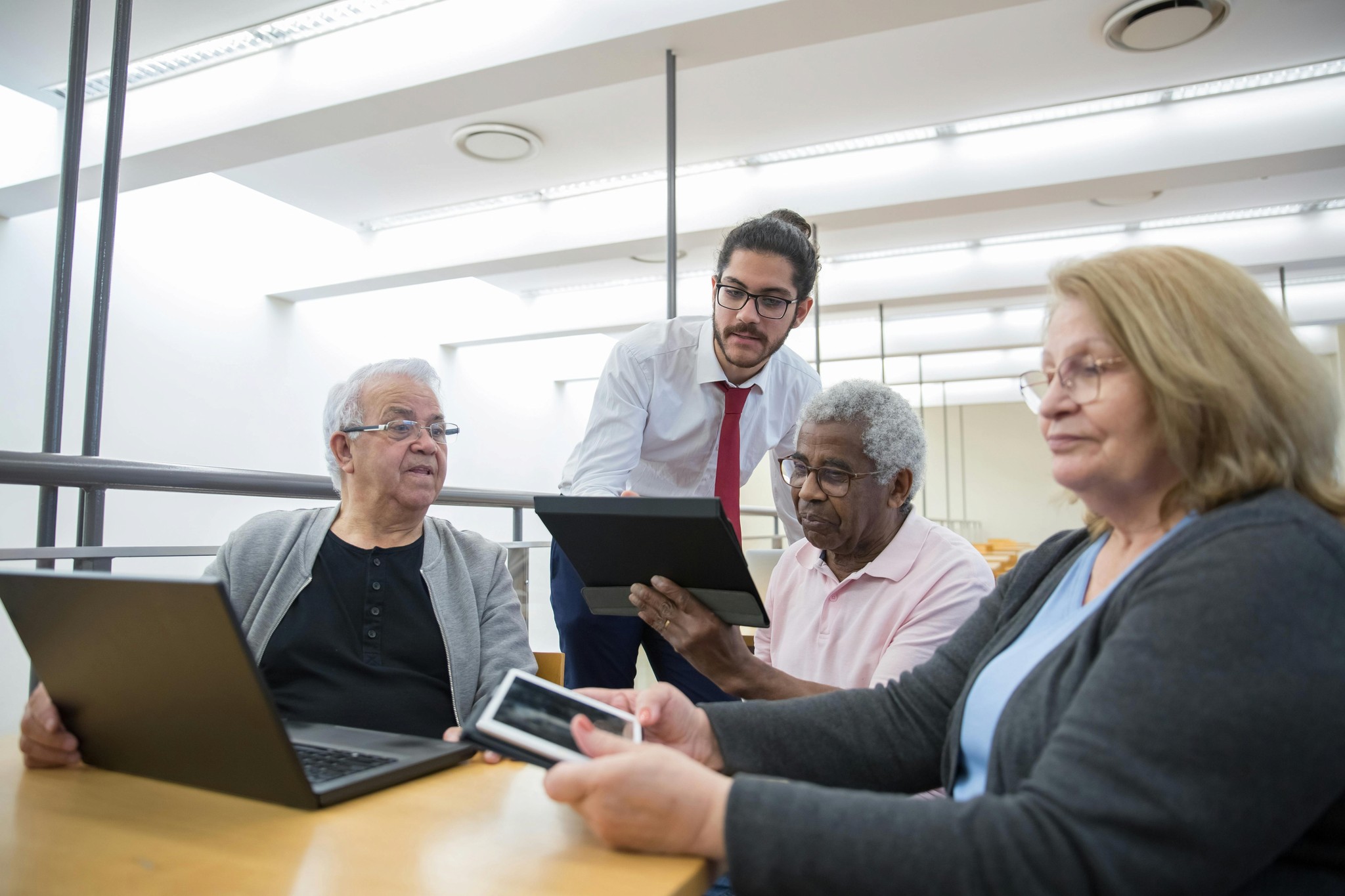 3 diverse senior citizens sit at a table lookign at their devices while a younger gentleman observes them