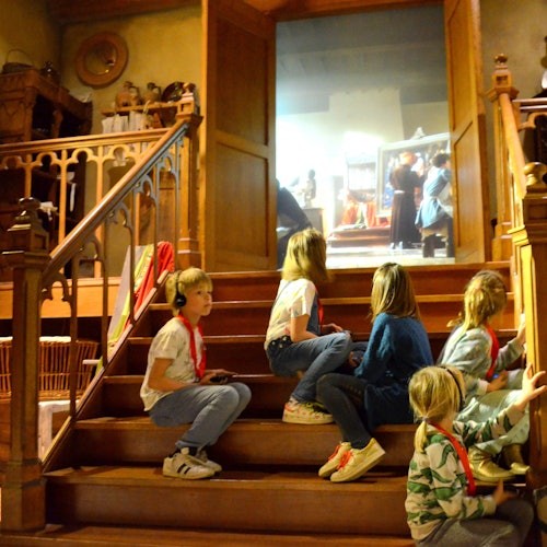 Négy gyerek ül egy díszes épület lépcsőjén, néhányan fejhallgatót viselnek. Egy nyitott ajtón keresztül néznek be.