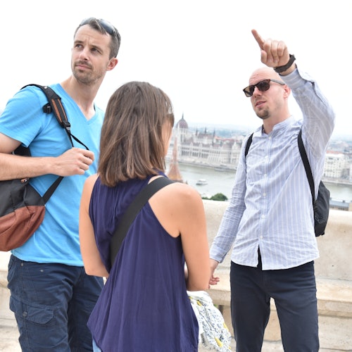 Three people with backpacks stand on a balcony, with one person pointing towards a distant cityscape.