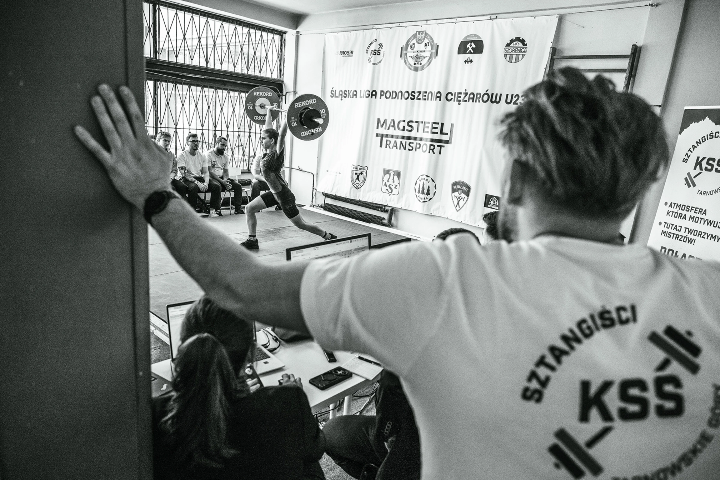 Czarno-białe zdjęcie z zawodów Śląskiej Ligi Podnoszenia Ciężarów U23 – zawodniczka podnosząca sztangę na pomoście.