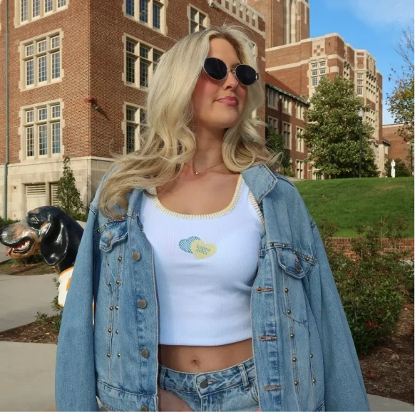 Outdoor campus photo of Blaire Latham wearing a white Kappa Delta tank with yellow trim and a denim jacket, posing near a brick university building.