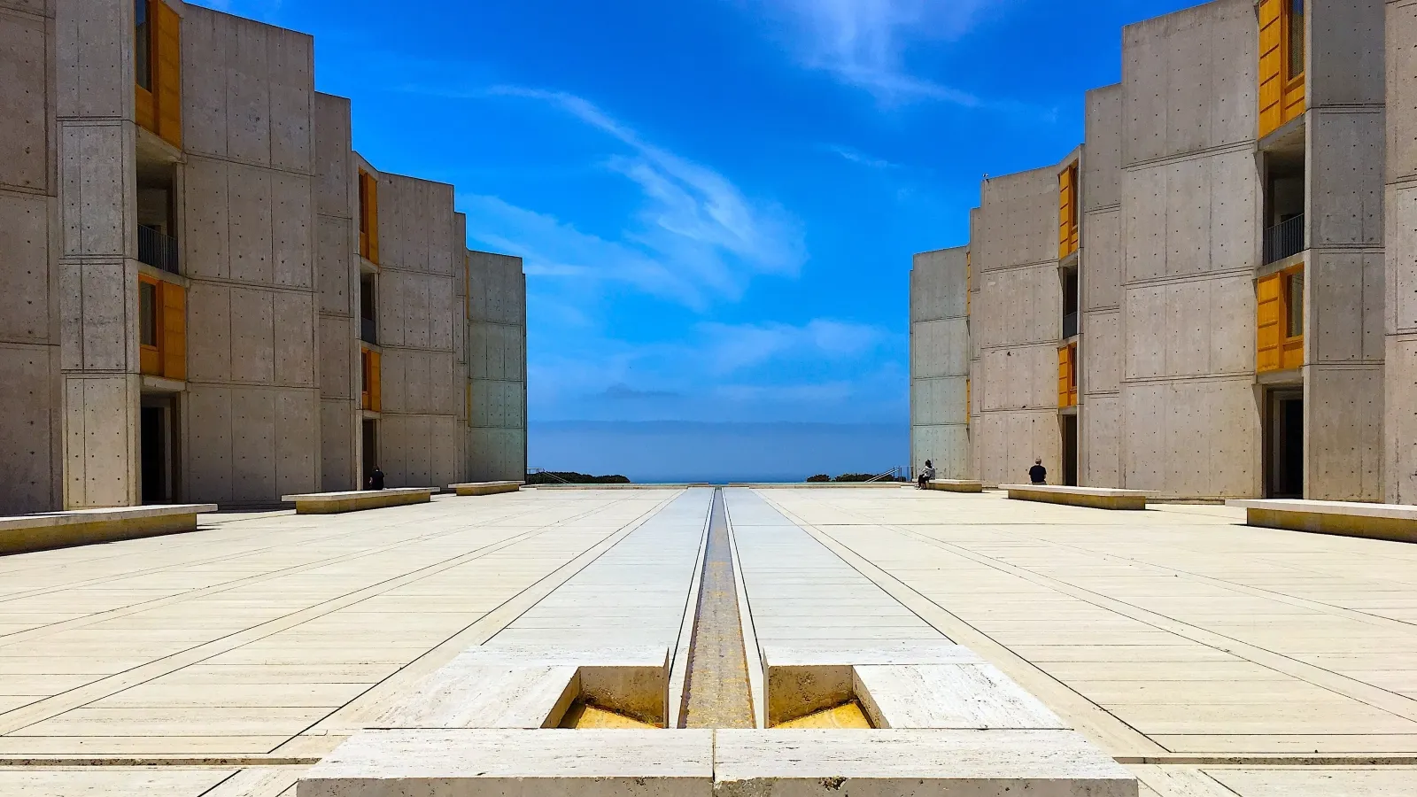 A symmetrical view of the Salk Institute's courtyard shows two concrete buildings framing a pathway that leads the eye towards the horizon, under a bright blue sky with wispy clouds.