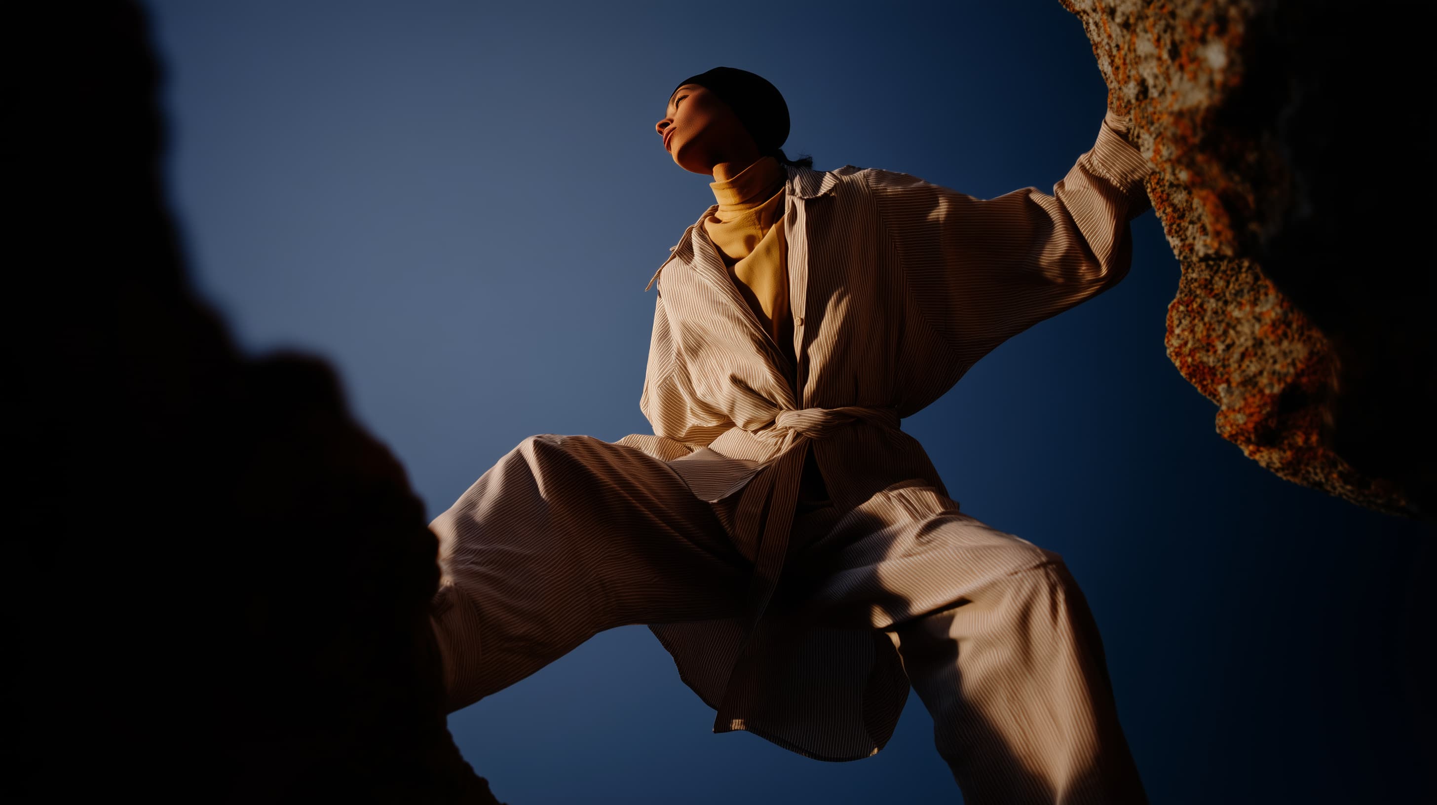 Low-angle editorial fashion shot of a person in neutral-toned clothing standing against a deep blue sky among rocks.