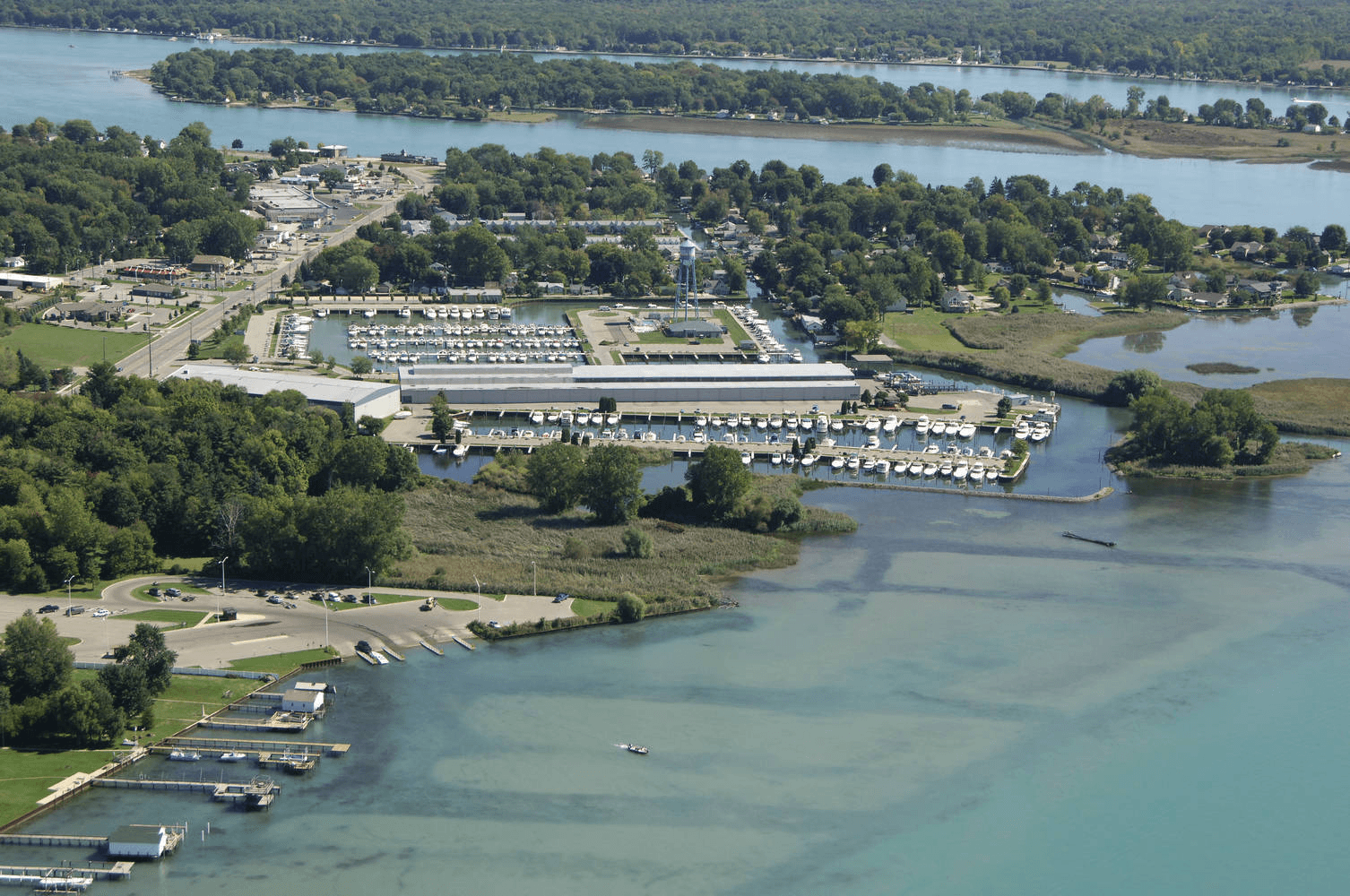 Aerial view of Algonac Harbour Club on the North Channel, rows of slips flanking a long indoor storage building, with a public launch ramp, the delta marshes and the St. Clair River beyond