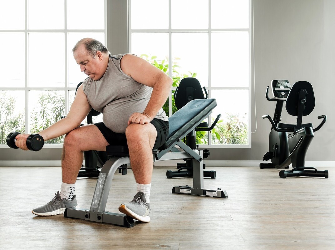 overweight older man doing beginner workouts for losing weight at the gym by using dumbbells for bicep curls