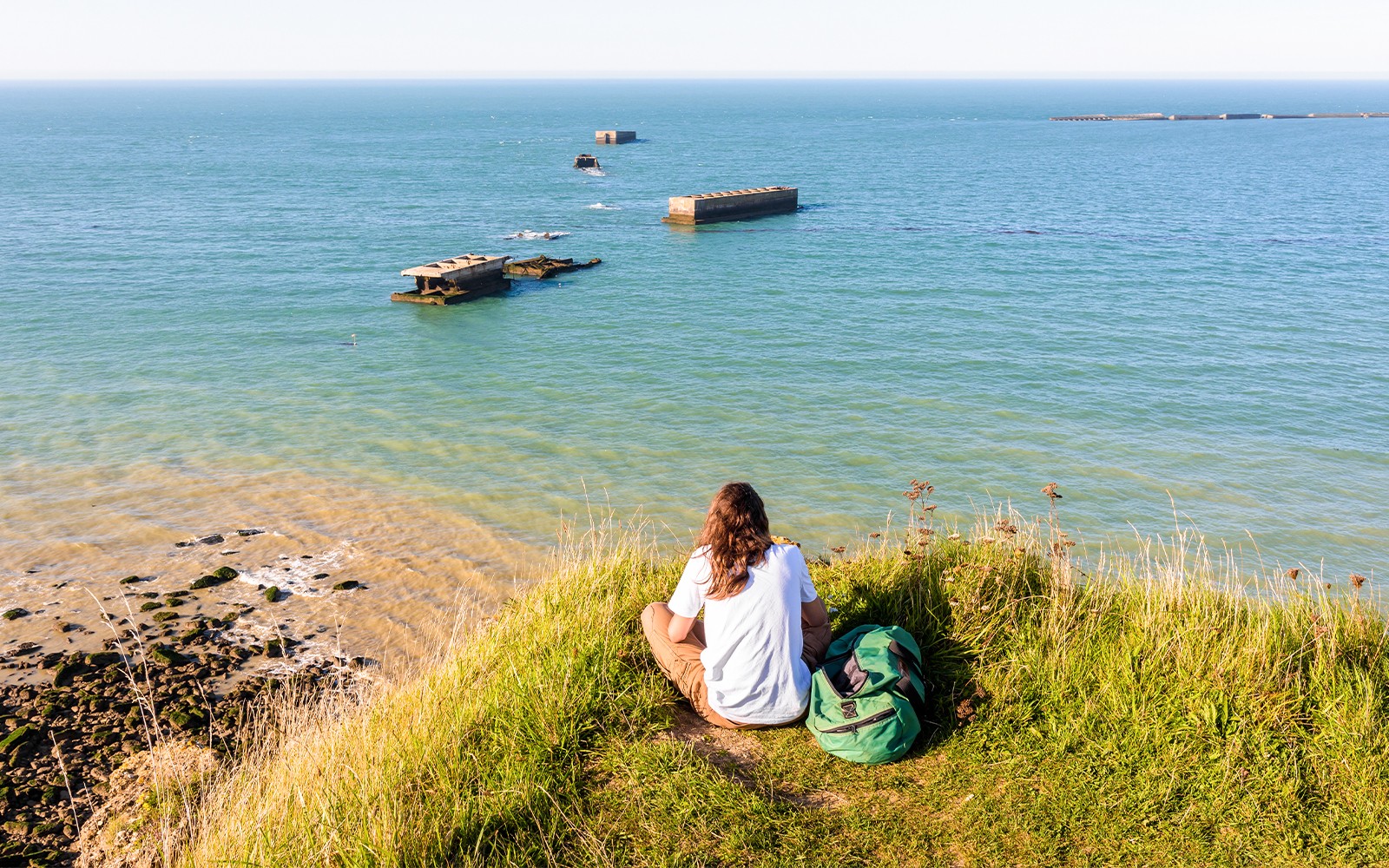 Person sitting on a grassy cliff overlooking the Normandy D-Day beaches with WWII remnants in the sea.