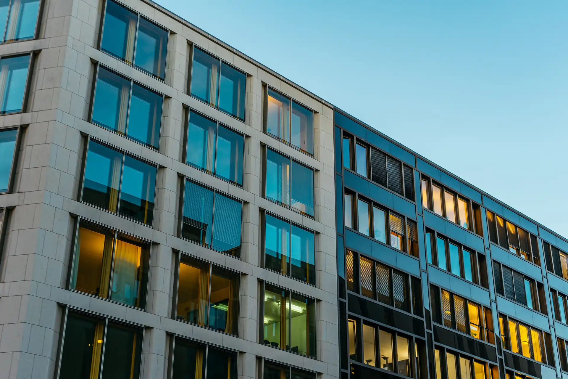 A low-angle view of the facade of a modern commercial building with a grid-like design. On the left, the structure is clad in light-colored stone blocks with bluish-tinted windows; on the right, the structure is dark, featuring windows that reflect a warm, yellowish interior light, set against a clear blue sky.
