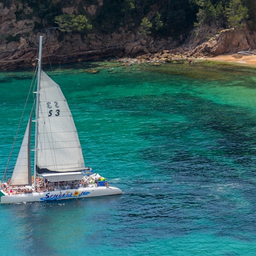 Voilier avec des personnes sur le pont glissant sur une eau turquoise claire près d'une plage rocheuse avec des arbres verts en arrière-plan.