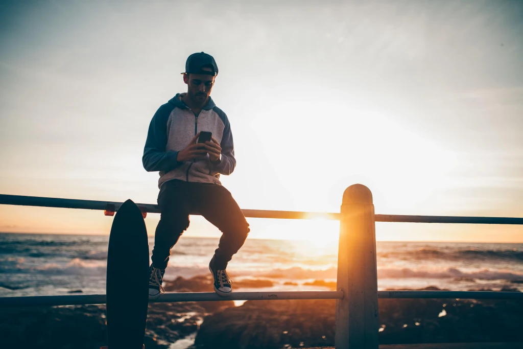 A young man on his phone at the beach with his back to a sunset