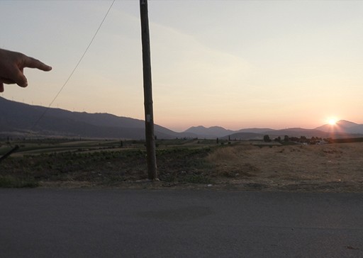 Χέρι δείχνει προς τον ορίζοντα σε ένα τοπίο της Δυτικής Αττικής, με τον ήλιο να δύει πίσω από τα βουνά.