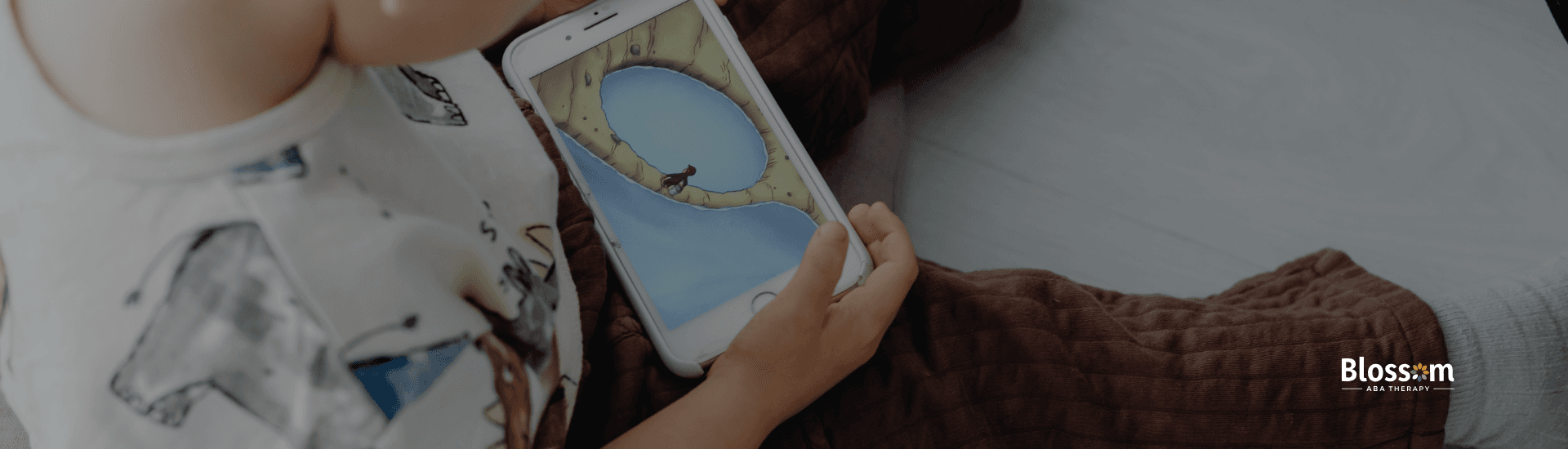 Close-up of a young child holding and looking at a smartphone while seated indoors, with an illustrated game or video on the screen; Blossom ABA Therapy logo visible in the corner.