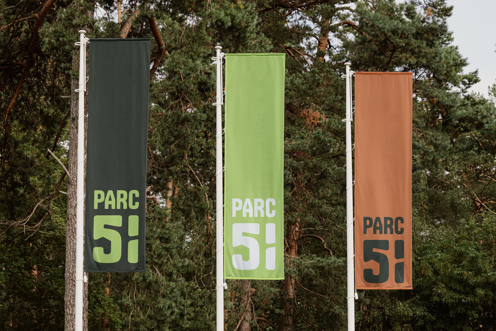 Drie hoge, verticale banier-vlaggen die wapperen voor een rij naaldbomen. De middelste vlag is opvallend groen met het witte Parc51-logo, geflankeerd door een donkergrijze en een roestbruine vlag.