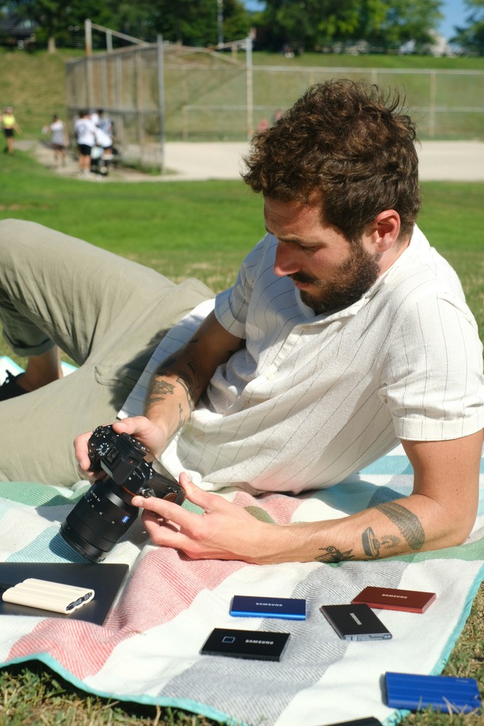 A bearded man with tattoos relaxes on a picnic blanket in a park, examining his camera. Surrounding him are various Samsung portable SSDs and a notebook, suggesting a tech-savvy photographer reviewing his equipment on a sunny day. A baseball field and other park-goers are visible in the background.