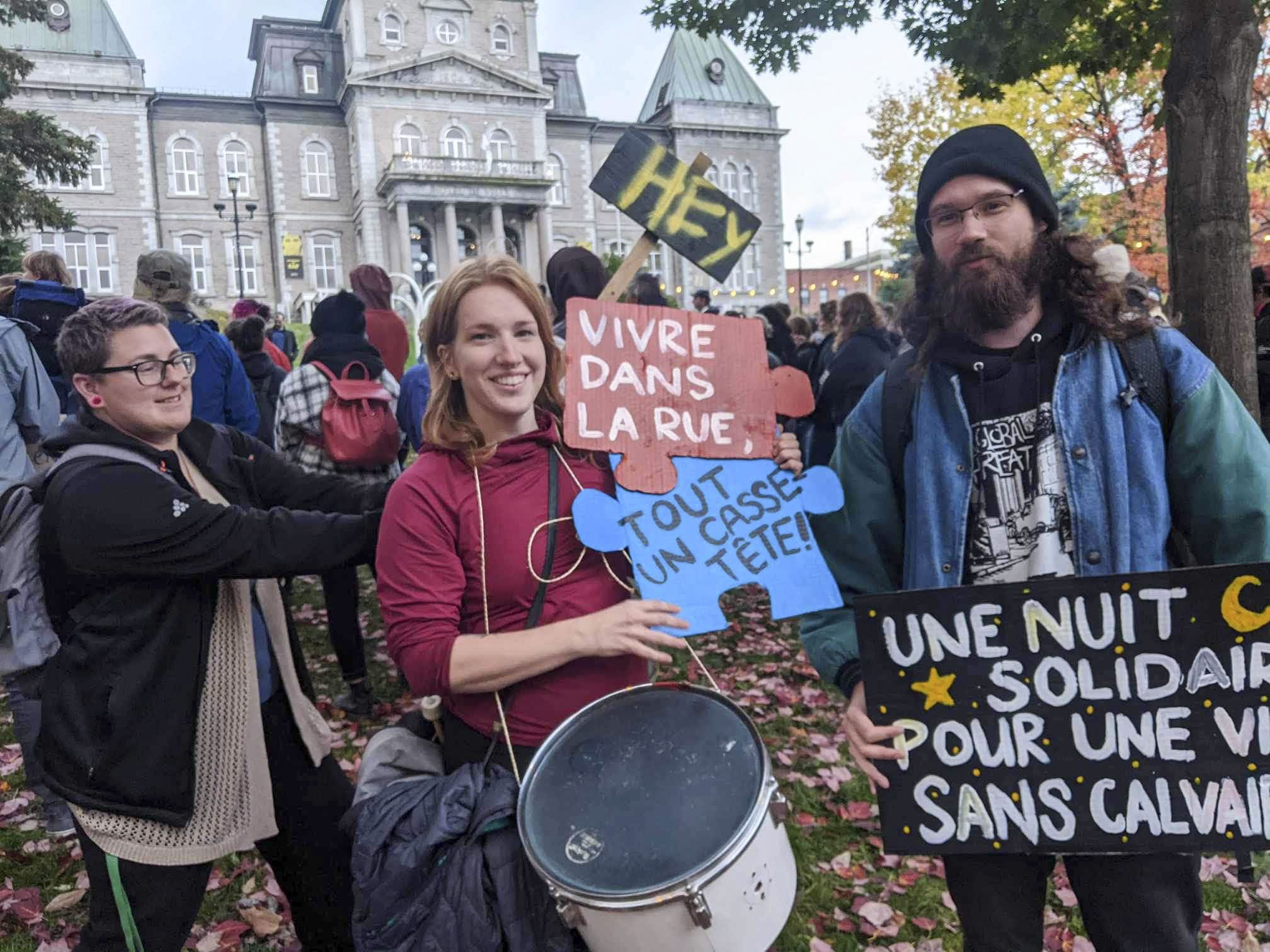 Deux personnes souriantes tenant des pancartes lors d'une manifestation, l'une avec un tambour, l'autre portant une affiche en soutien aux personnes en situation d’itinérance.
