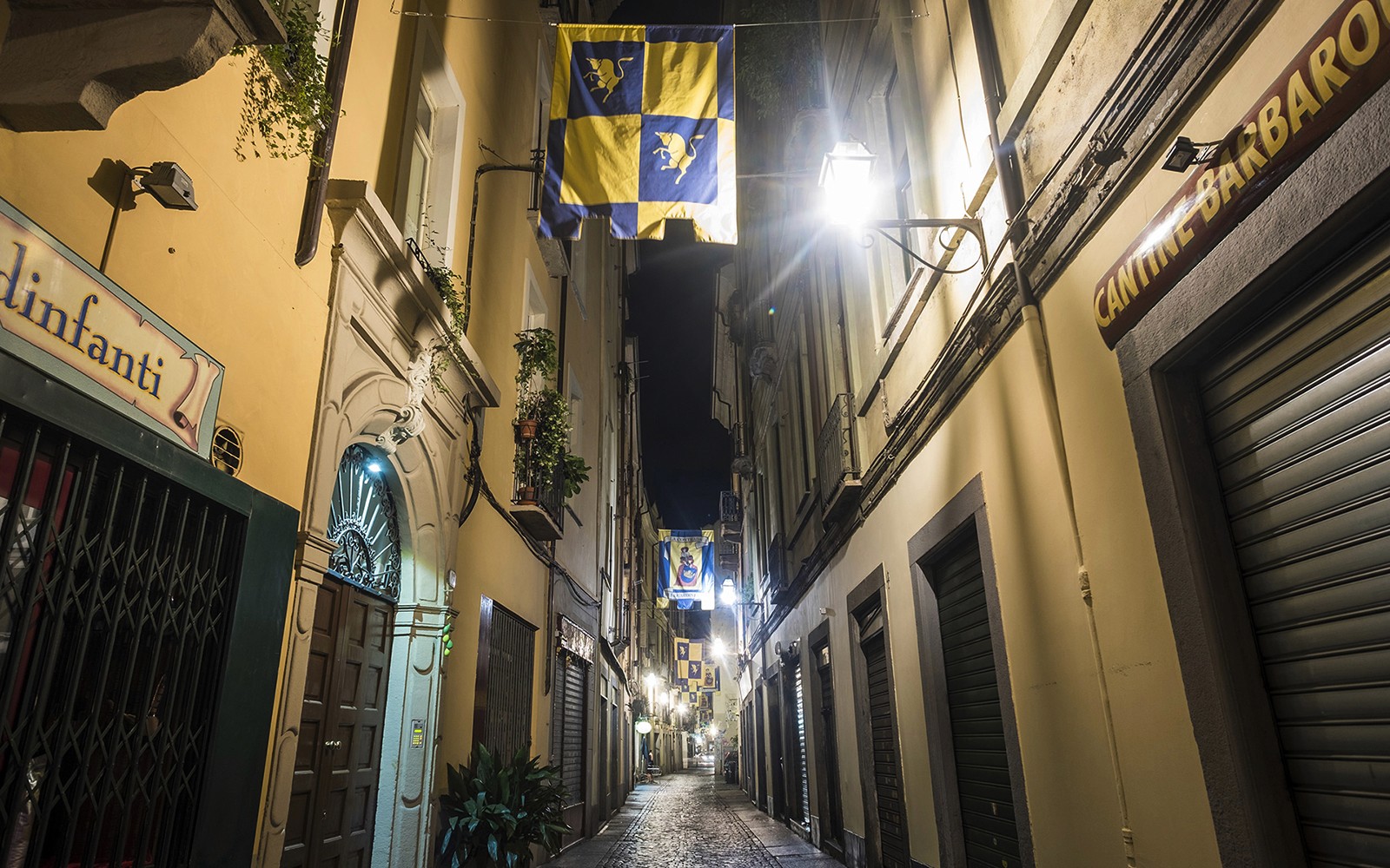 Narrow cobblestone street in Torino at night with flags and historic buildings.