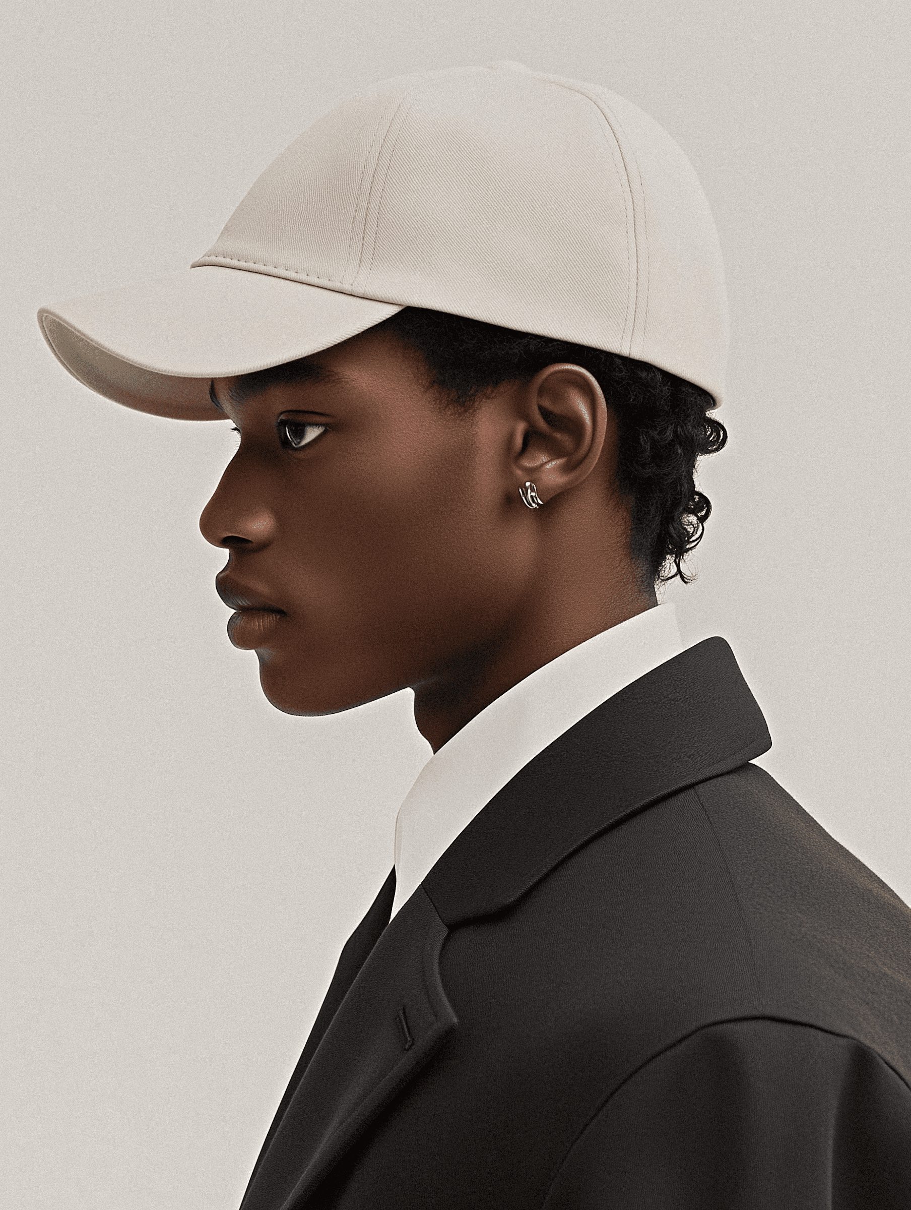 Stylish side profile of a man in a cream cap, black coat, and white shirt against a minimalist gray background.