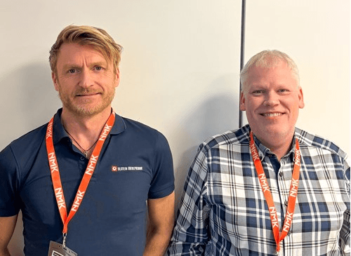 Two men stand side by side, smiling, in a well-lit room. One wears a dark shirt, and the other a plaid shirt.
