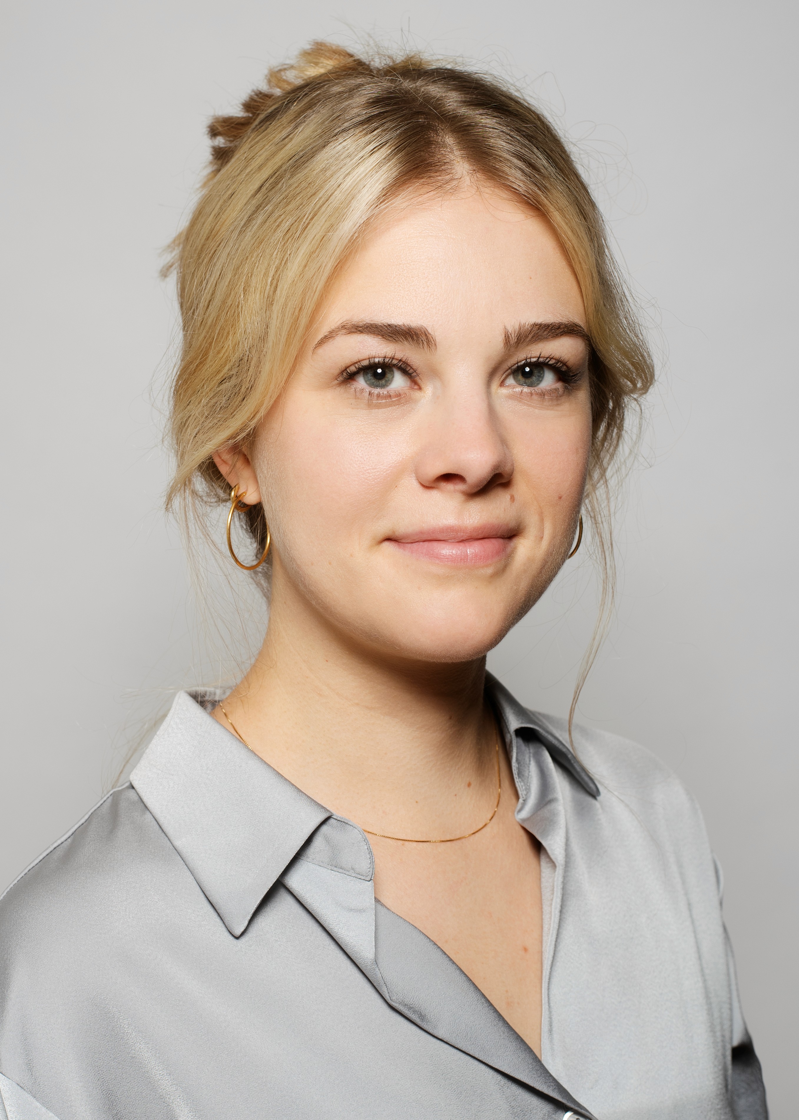 Eine junge Frau mit blondem Haar, die ein hellgraues Hemd trägt, lächelt in die Kamera vor einem neutralen Hintergrund.