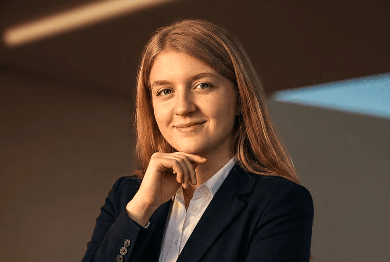 Woman with a short haircut smiling confidently, wearing a blazer and standing in warm natural light with modern architecture behind her.