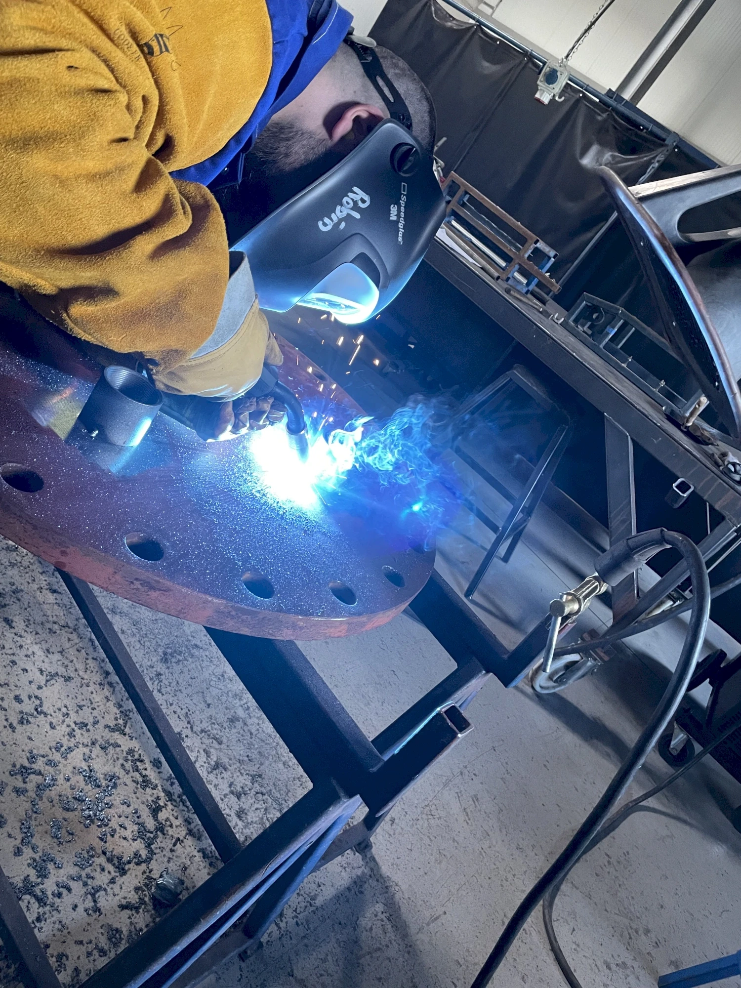 A welder wearing a mask and gloves welds a metal object in an industrial setting, with bright sparks visible.