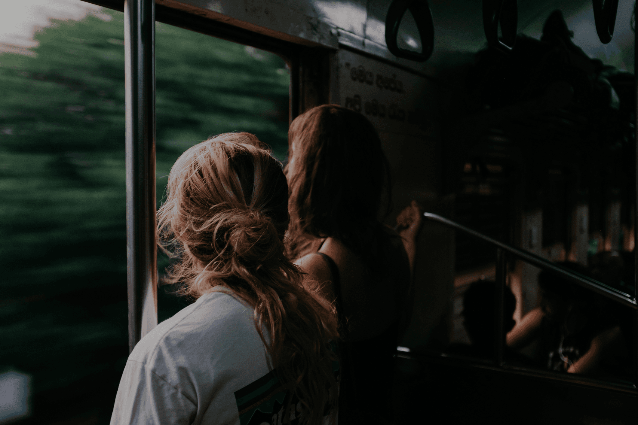 Two women standing by a train window, watching blurred green landscape pass outside, capturing a cinematic travel moment with natural light and motion blur.