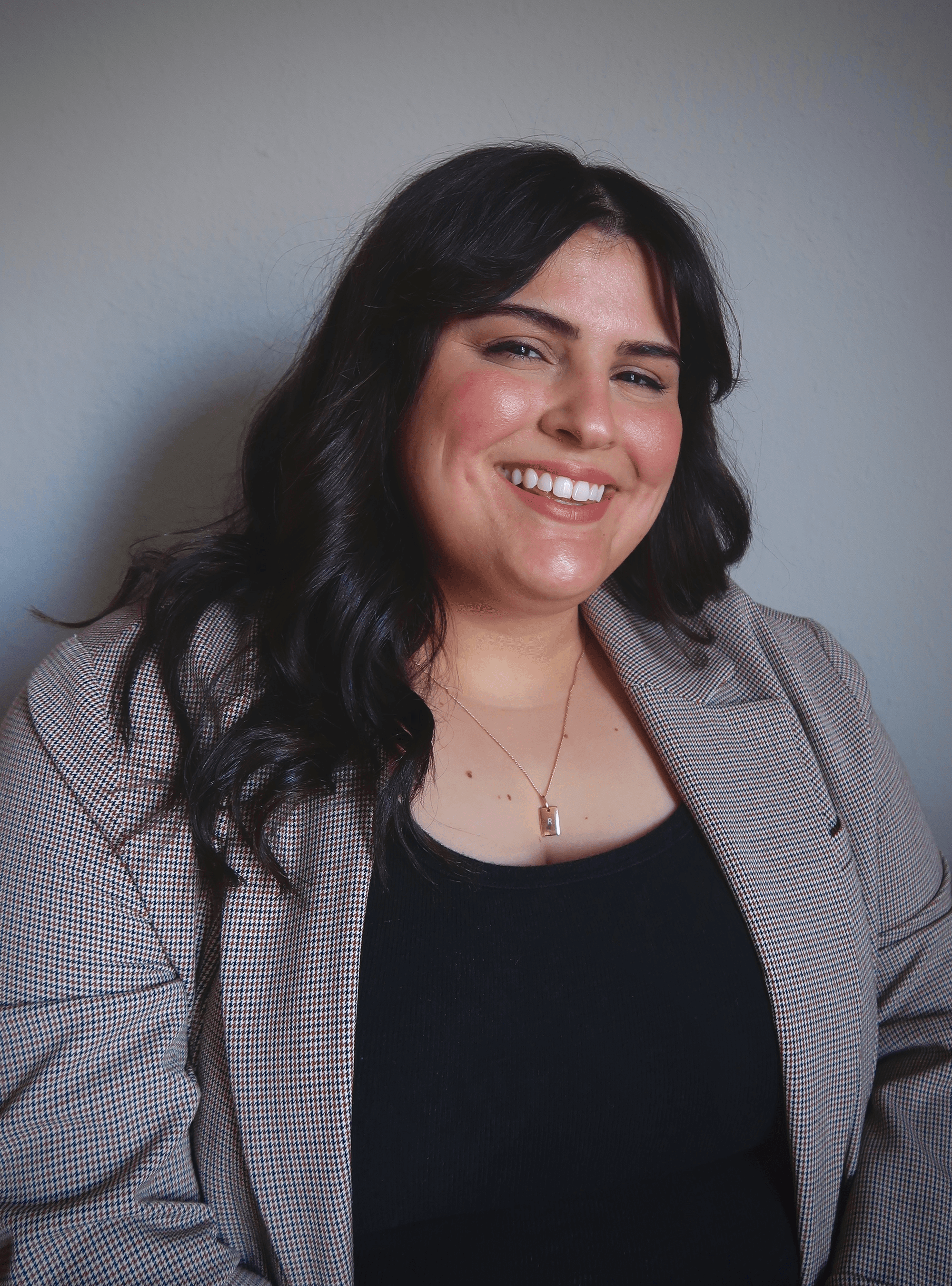 Smiling woman with long dark hair wearing a black top and gray blazer against a neutral background.